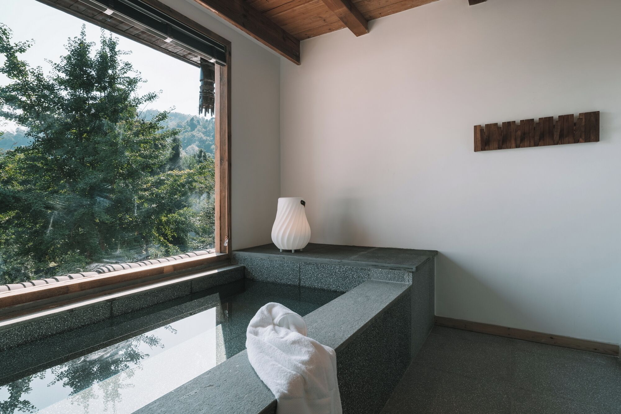 Un baño moderno con encimera de piedra, una toalla blanca y vistas a un bosque a través de un ventanal amplio.