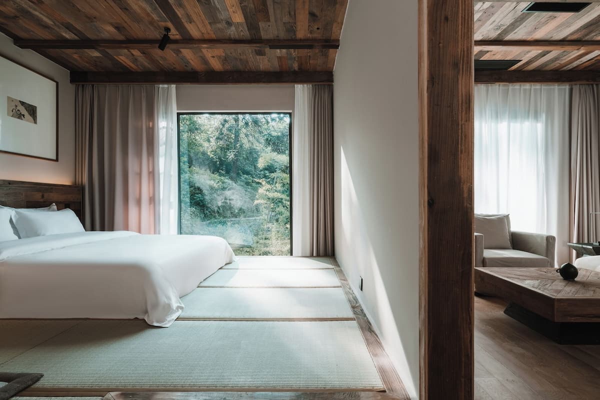 Une chambre d’hôtel avec un grand lit, une vue sur la verdure, un plafond en bois et un petit coin salon.