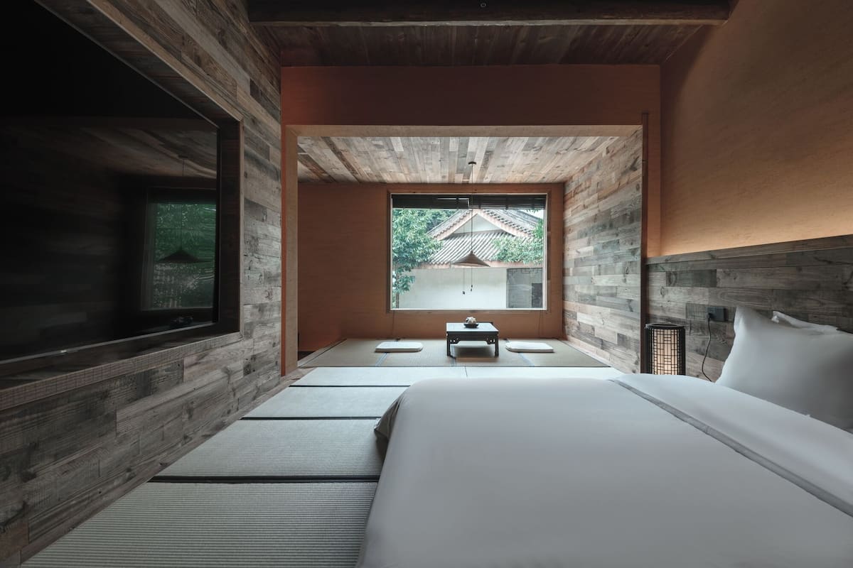 Une chambre au décor minimaliste, avec un grand lit, une petite table et une fenêtre donnant sur un bâtiment traditionnel.