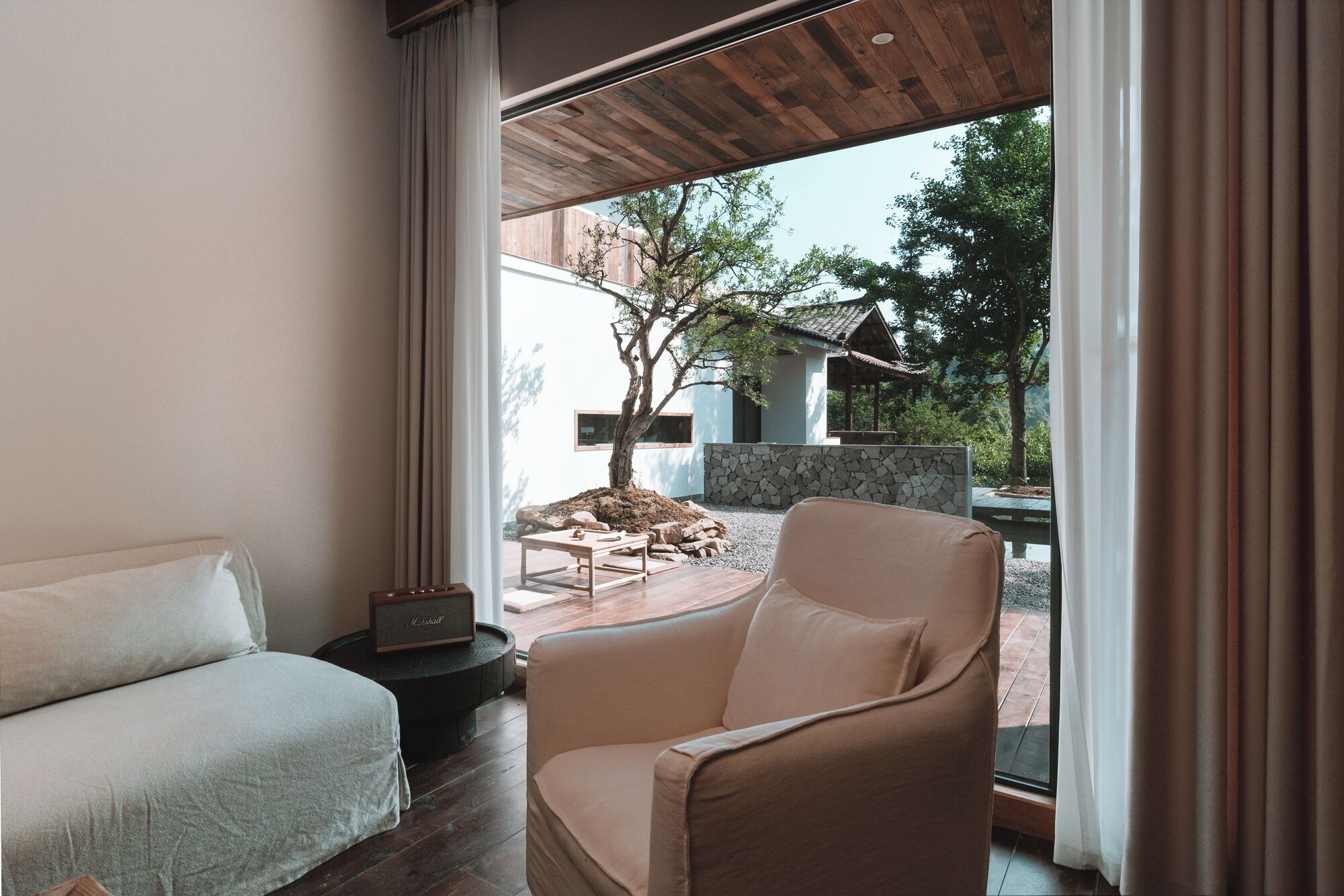 A modern room with a bed, a chair, and a view of an outdoor area with a tree and a stone wall.
