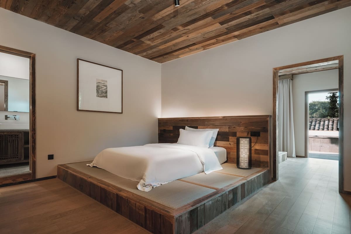 Une chambre à coucher avec une tête de lit en bois, un lit recouvert de draps blancs, un tableau encadré accroché au mur et une vue sur l’extérieur par une porte ouverte.