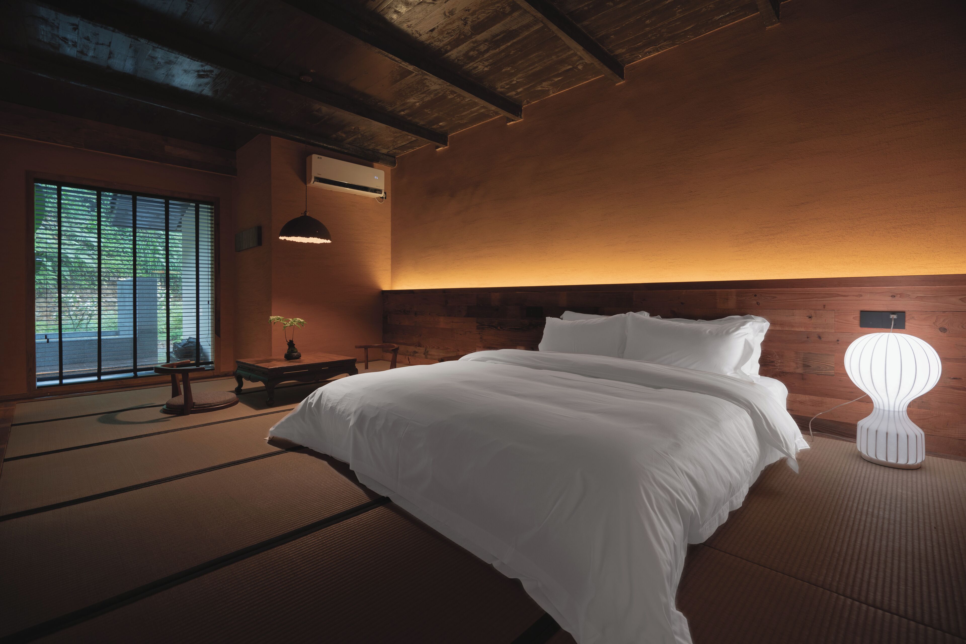 Une chambre de style japonais traditionnel, avec un sol recouvert de tatamis, un grand lit, une table de chevet et une fenêtre équipée de stores.