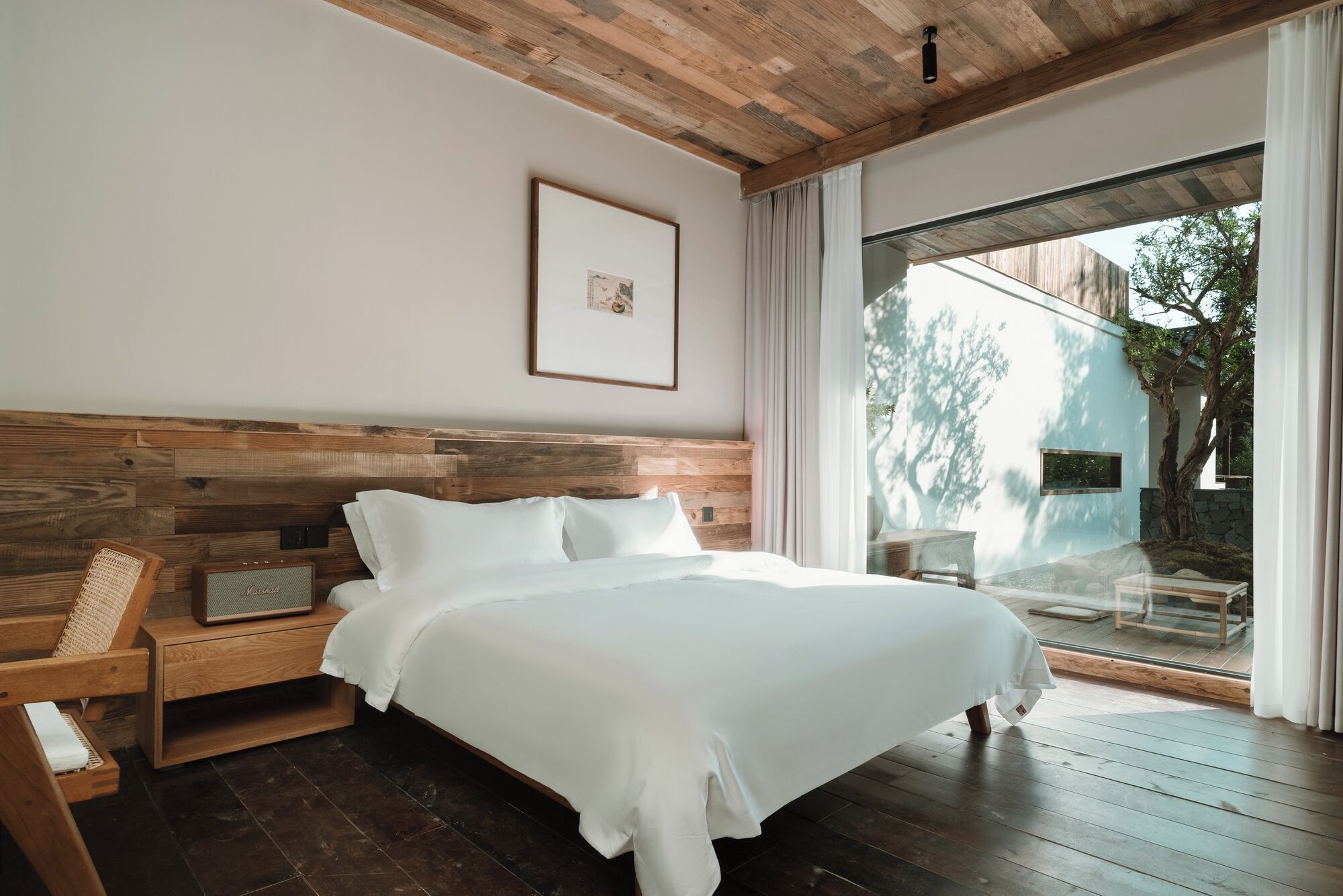 Un dormitorio con cabecera de madera, cama con ropa de cama blanca, mesita de noche con lámpara y vistas al exterior a través de un ventanal.