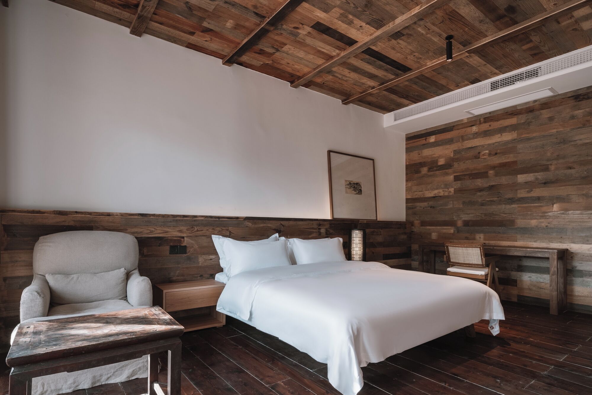Un dormitorio con techo de madera, cama con ropa de cama blanca, una silla y una mesita.