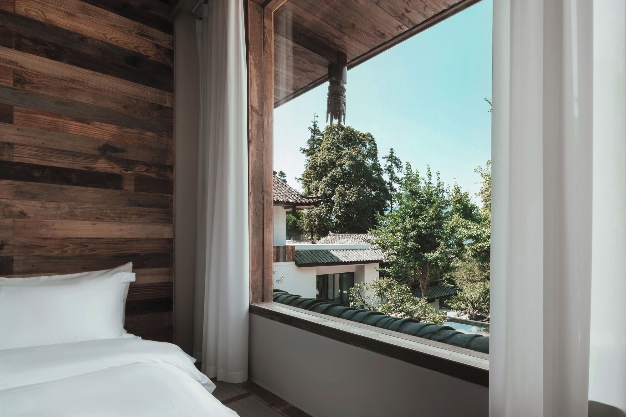 Una habitación con pared de madera, una cama con ropa blanca y una ventana con vista a un edificio tradicional y árboles.