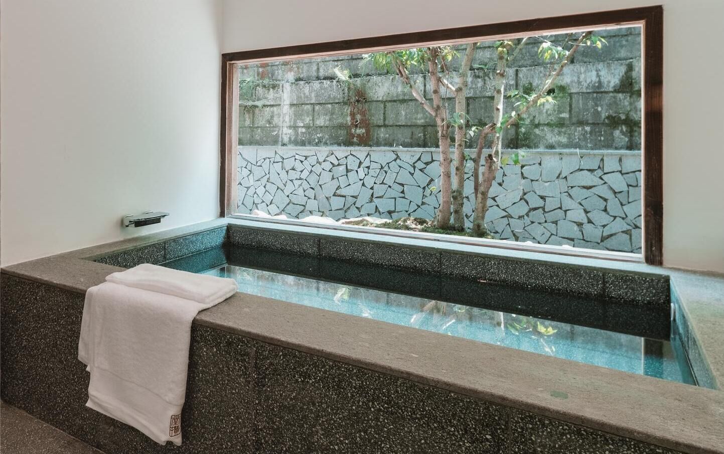 A spa area with a stone bathtub, a window with a view of trees and a stone wall, and a towel on the edge of the tub.