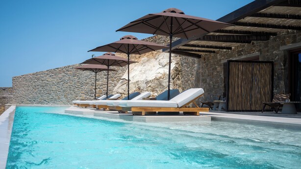 Una piscina con sillones de descanso y sombrillas, un muro de piedra y una terraza de madera.