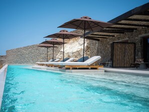 A pool with lounge chairs and umbrellas, a stone wall, and a wooden deck. - Petrablu Mykonos (Mykonos)