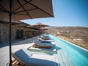 A poolside area with lounge chairs and umbrellas, stone walls, and a view of the landscape. - Petrablu Mykonos (Mykonos)