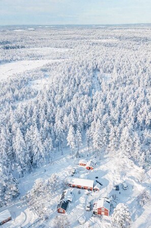 Un paysage enneigé avec un ensemble de bâtiments rouges, probablement un chalet ou des cabanes, entouré d’une forêt dense.