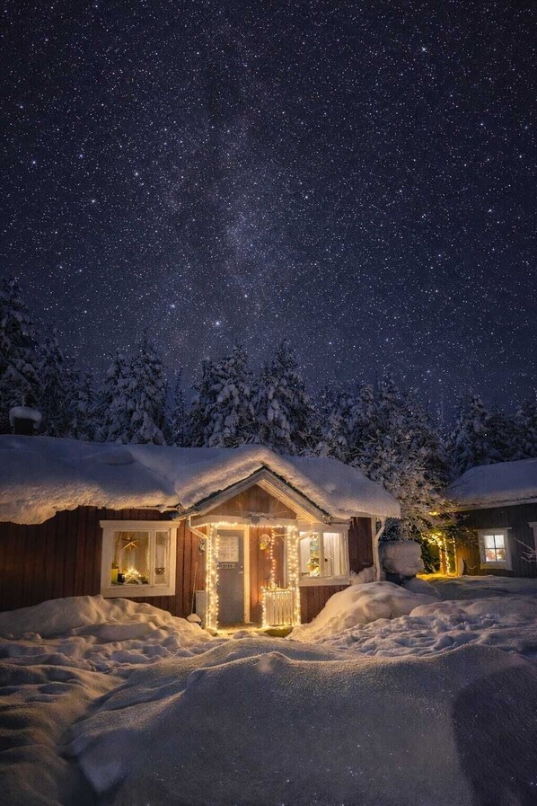 Una cabaña nevada con la entrada iluminada, rodeada de nieve bajo un cielo estrellado.