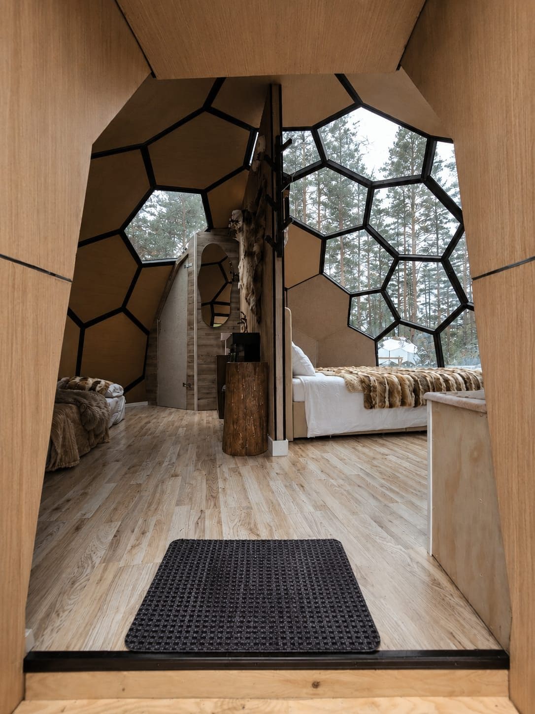 A hexagonal room with a bed, a wooden floor, and a view of trees through a hexagonal window.