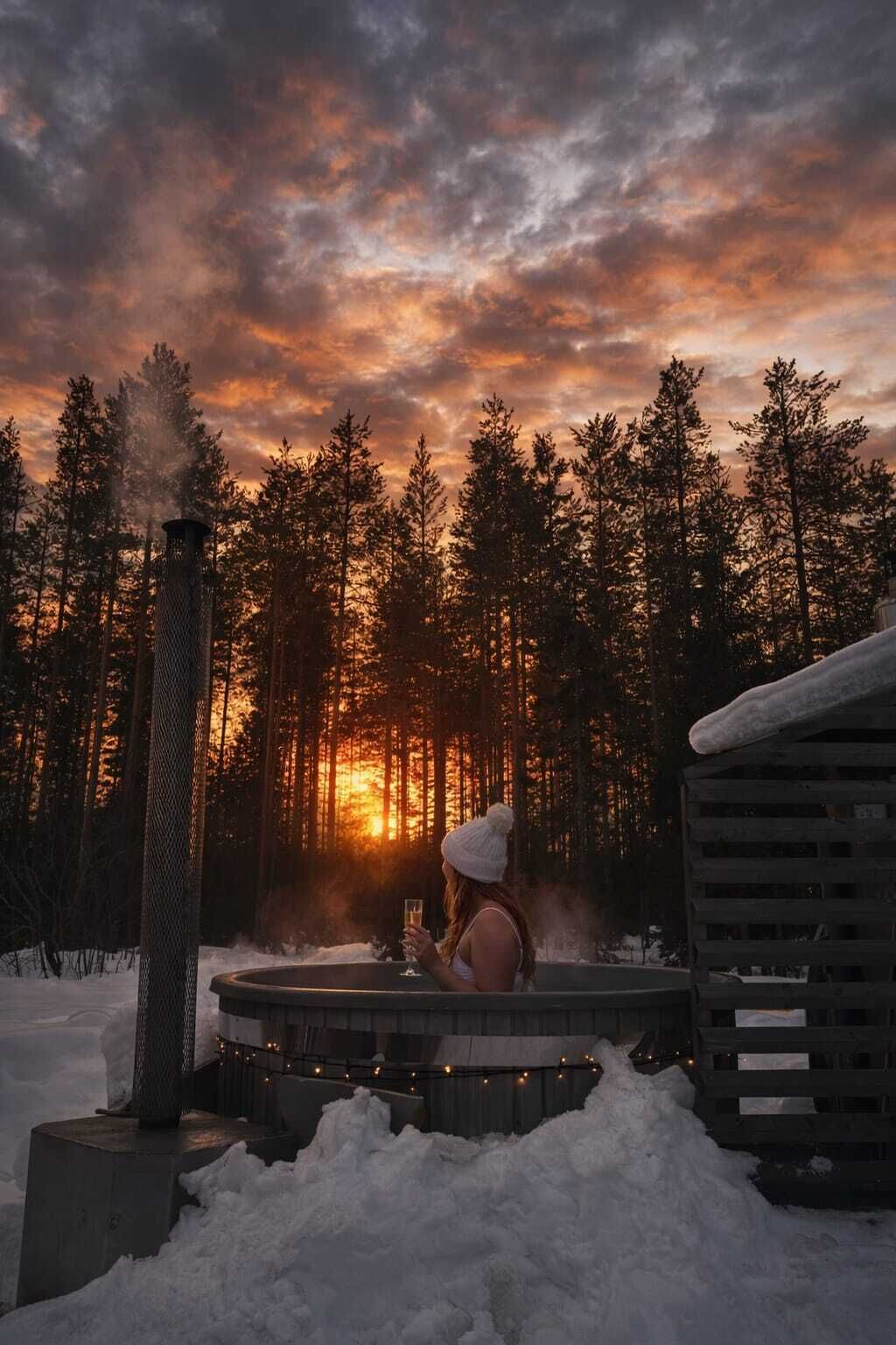 A hot tub surrounded by snow, a wooden cabin, and a forest with a sunset in the background.