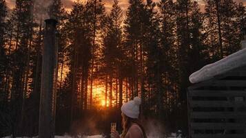A hot tub surrounded by snow, a wooden cabin, and a forest with a sunset in the background.