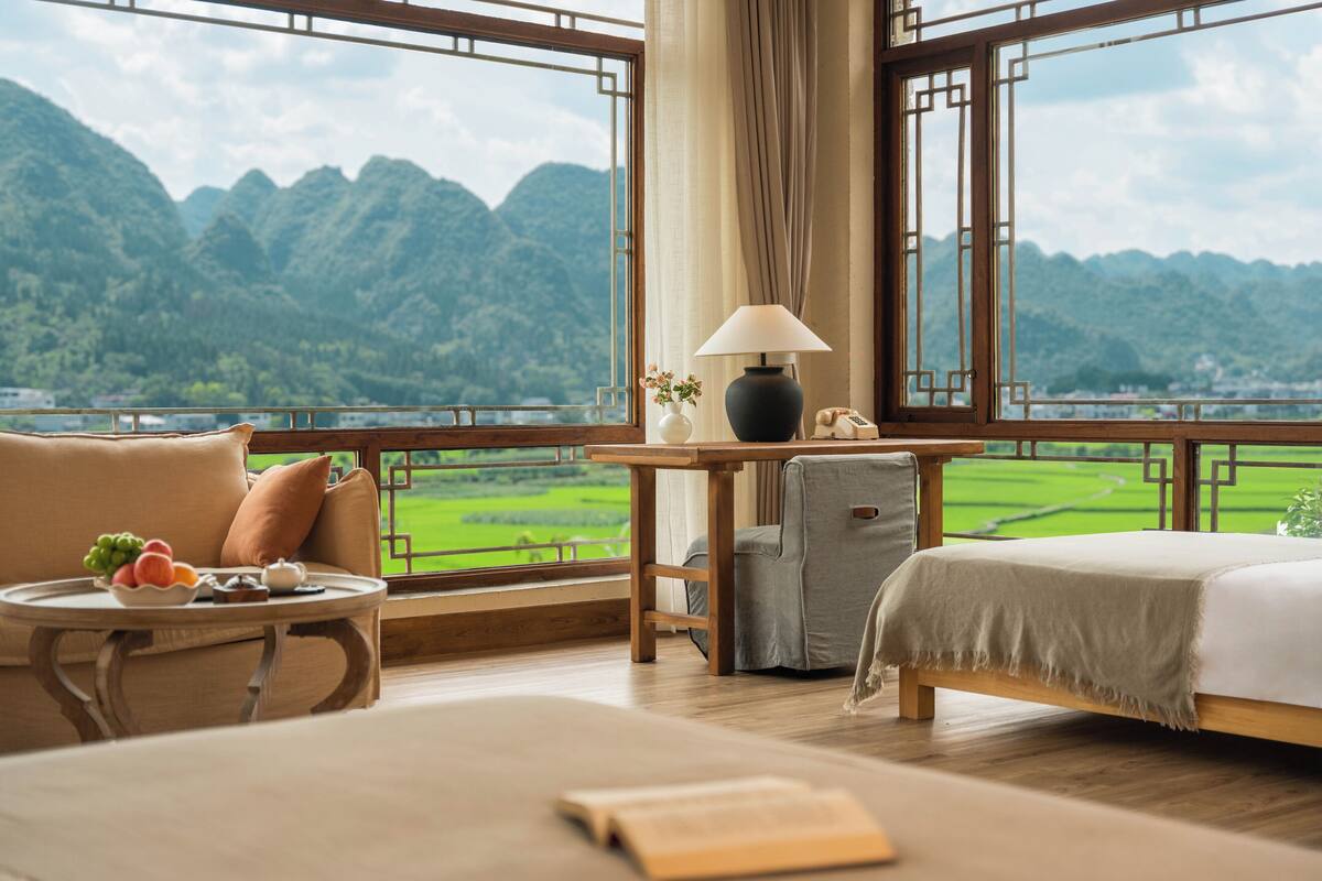 Ein Zimmer mit einem Bett, einem Tisch mit Lampe, einem Sessel und Bergblick durch große Fenster.
