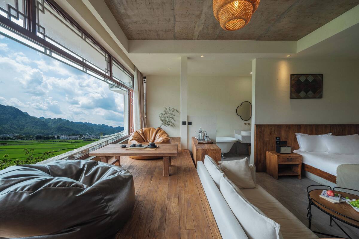Ein modernes Zimmer mit Holzboden, einem großen Fenster mit Blick auf grüne Felder und Berge, einem Bett mit weißer Bettwäsche, einem Nachttisch aus Holz und einem kleinen Tischchen mit einem Radio.
