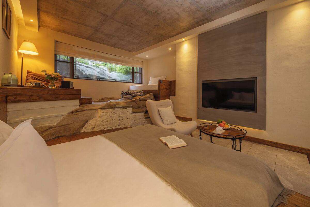 Ein modernes Schlafzimmer mit einem großen Bett, einer Sitzecke mit einem Sessel und einem kleinen Tisch, einem an der Wand montierten Fernseher und einer Wand mit Steinoptik.