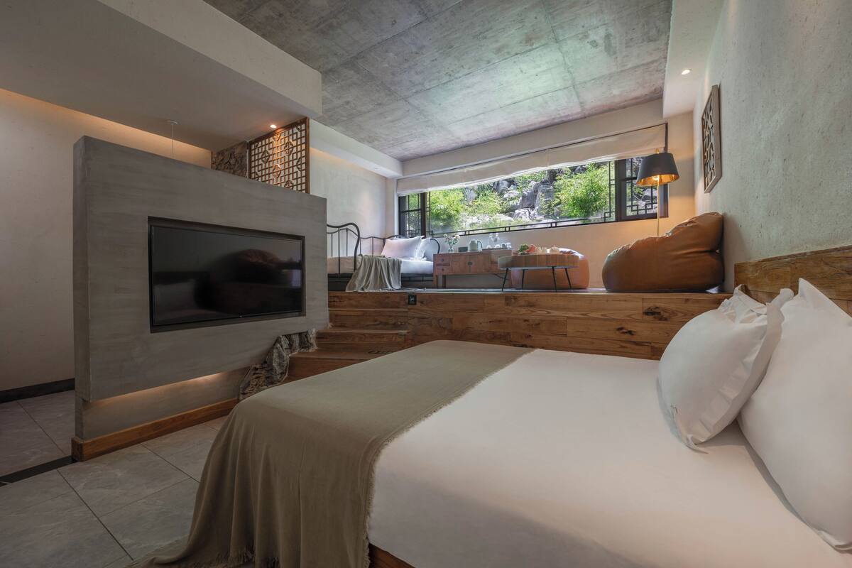 Ein modernes Schlafzimmer mit einem großen Bett, einem an der Wand montierten Fernseher, einer Holztreppe und Blick ins Grüne.