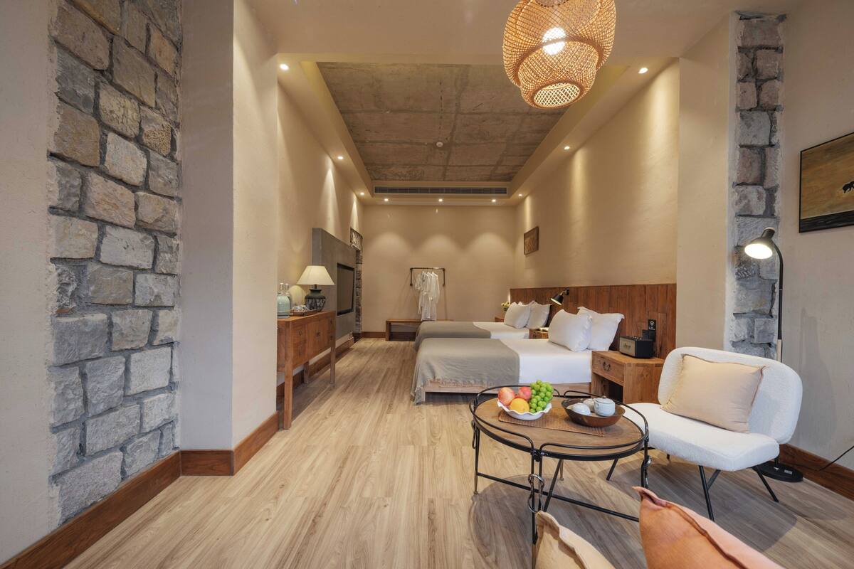 Ein Hotelzimmer mit Wandverkleidung aus Stein, Holzboden, einem Bett mit weißer Bettwäsche, einem Nachttisch mit Lampe, einem kleinen runden Tisch mit Obst und einem weißen Sessel.