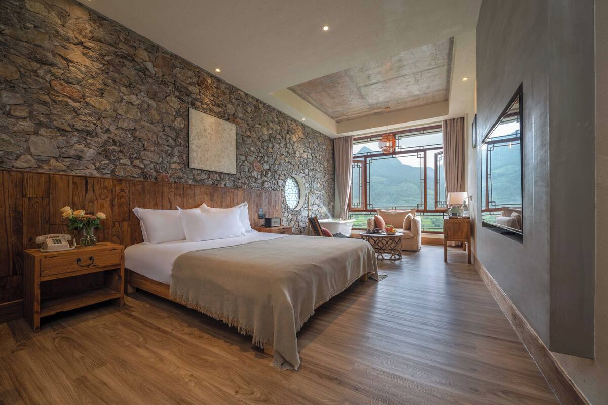 Ein geräumiges Schlafzimmer mit Steinswand, Holz-Kopfteil und einem großen Bett. Es gibt eine Sitzecke mit Blick auf die Berge.