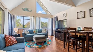 A spacious living room with a high ceiling, a large window, a dining area with a wooden table and chairs, and a television on a cabinet.