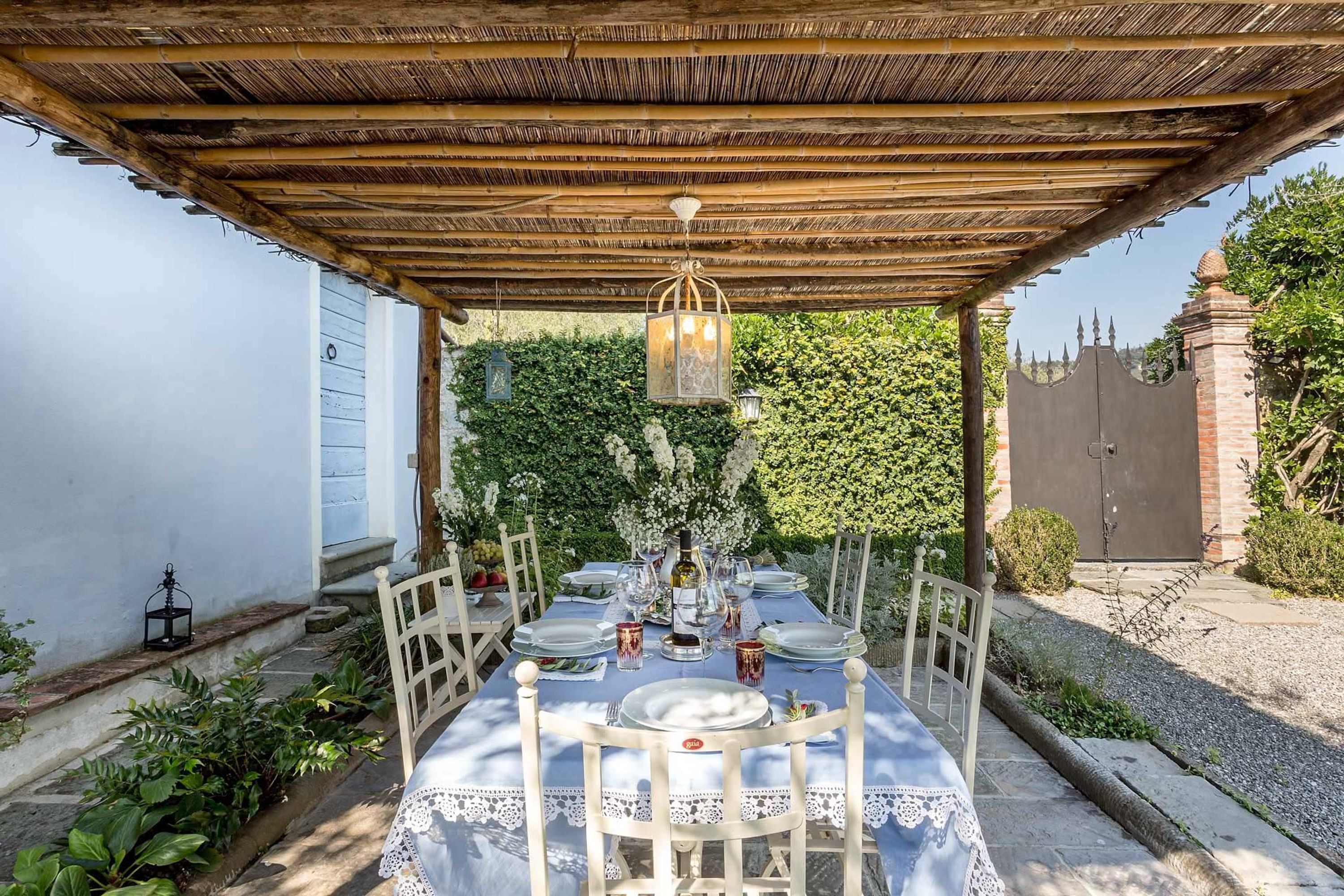 Un espace de restauration en plein air avec une table dressée pour quatre personnes, située sous une pergola en bois, entourée de verdure et d’un mur de briques.