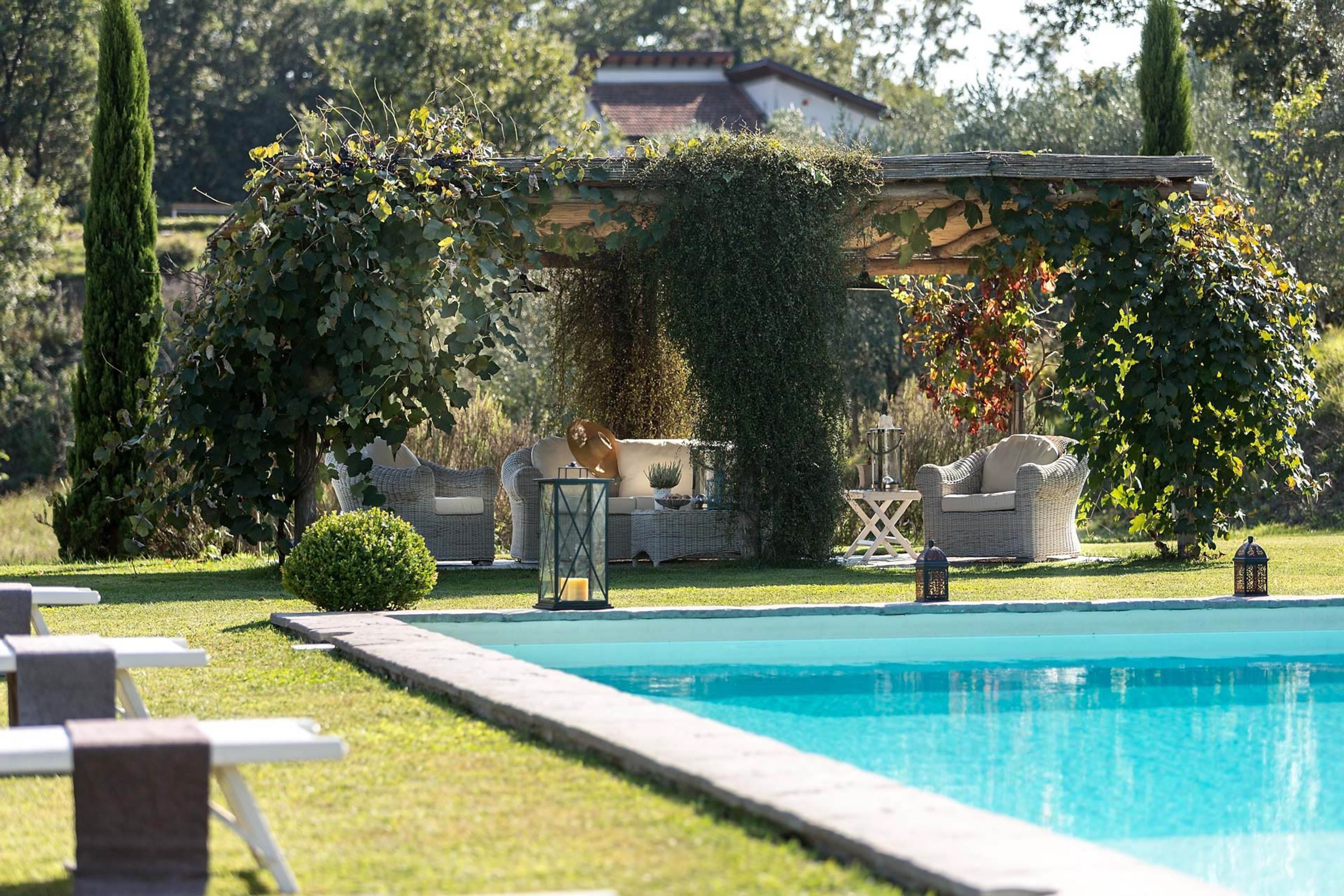 Zona de piscina con sillones de descanso, una pérgola cubierta de vegetación y un edificio al fondo.