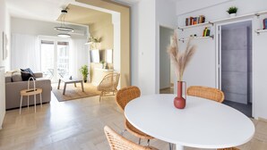 A modern living room with a round white table, wicker chairs, a sofa, and a coffee table. There is a bookshelf with books and a potted plant on the wall. - Santorini-2-Bedroom Apt in Nea Smyrni (Nea Smirni)