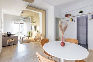 A modern living room with a round white table, wicker chairs, a sofa, and a coffee table. There is a bookshelf with books and a potted plant on the wall.