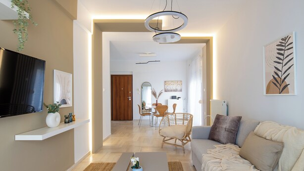 A modern living room with a sofa, coffee table, and a TV on a wall. There is a dining area with chairs and a table in the background. - Santorini-2-Bedroom Apt in Nea Smyrni (Nea Smirni)