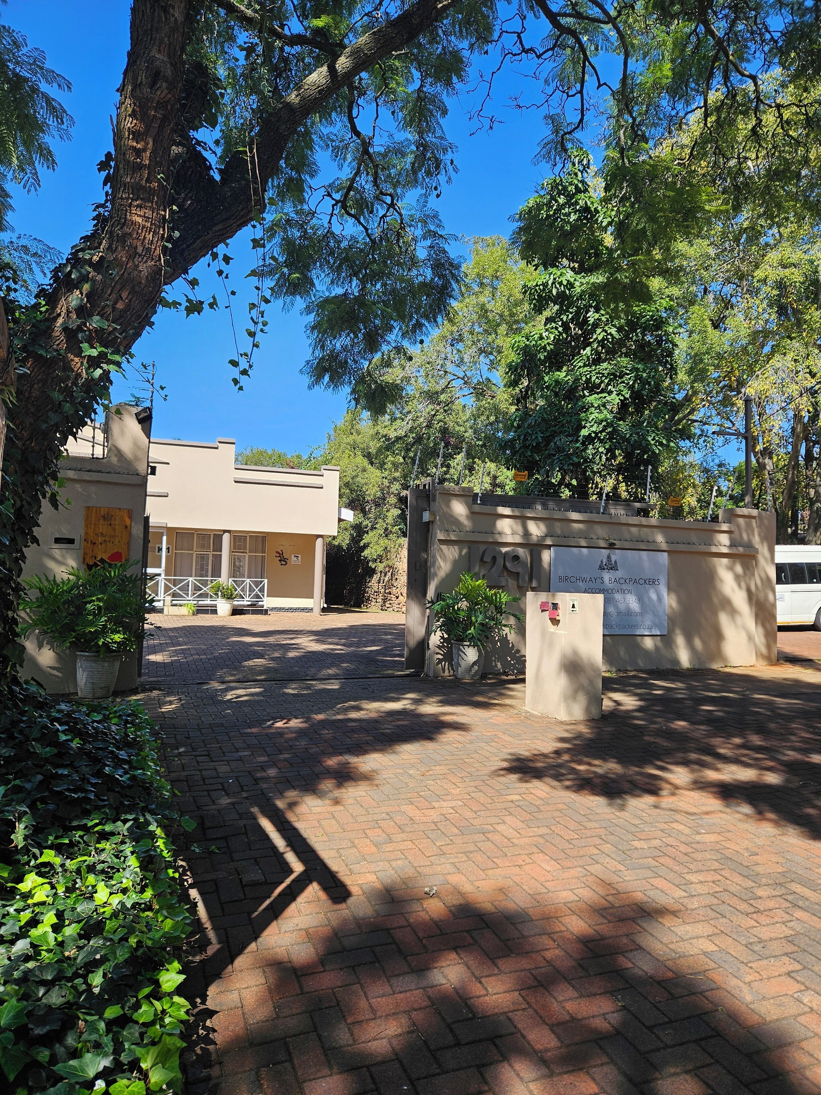 Une allée dallée avec un panneau indiquant « Birchway's Backpackers Accommodation », un bâtiment à la façade beige et un arbre aux feuilles vertes.