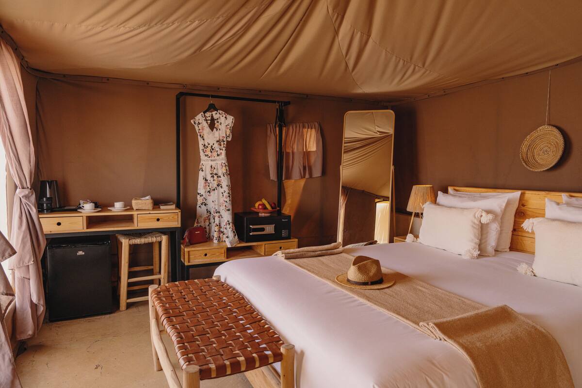 A bedroom with a bed, a desk, a chair, a small refrigerator, and a mirror.