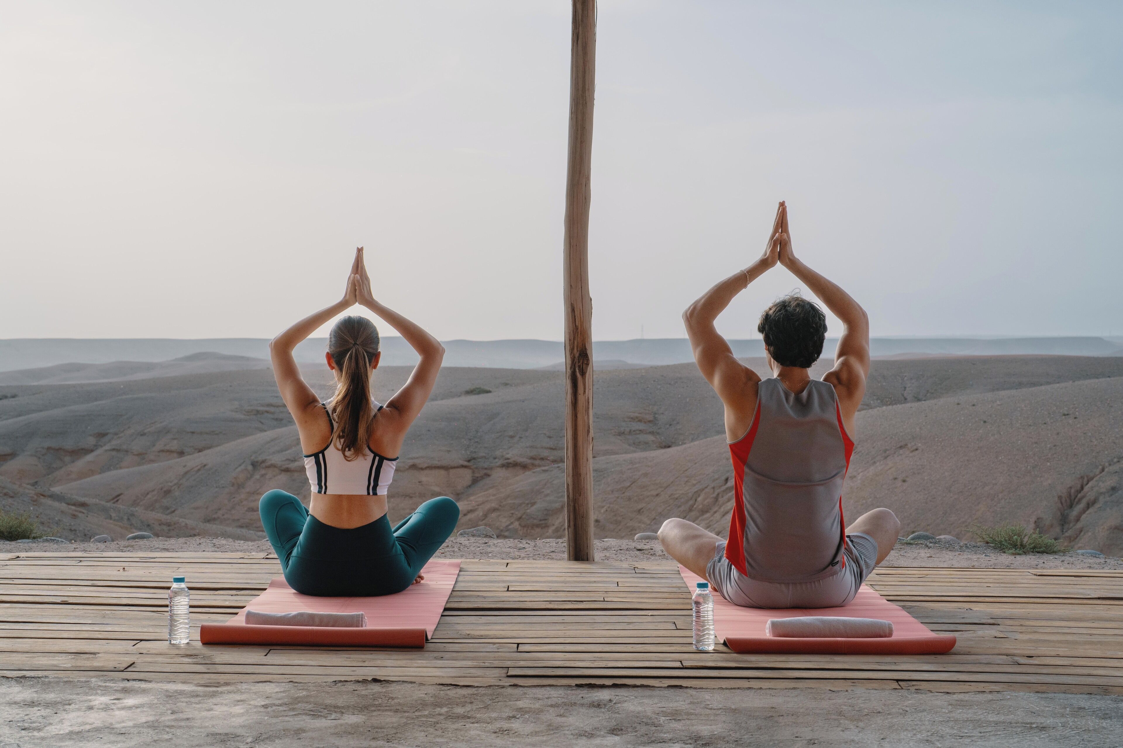 Two individuals practicing yoga on mats in an outdoor setting with a wooden deck and a mountainous backdrop.