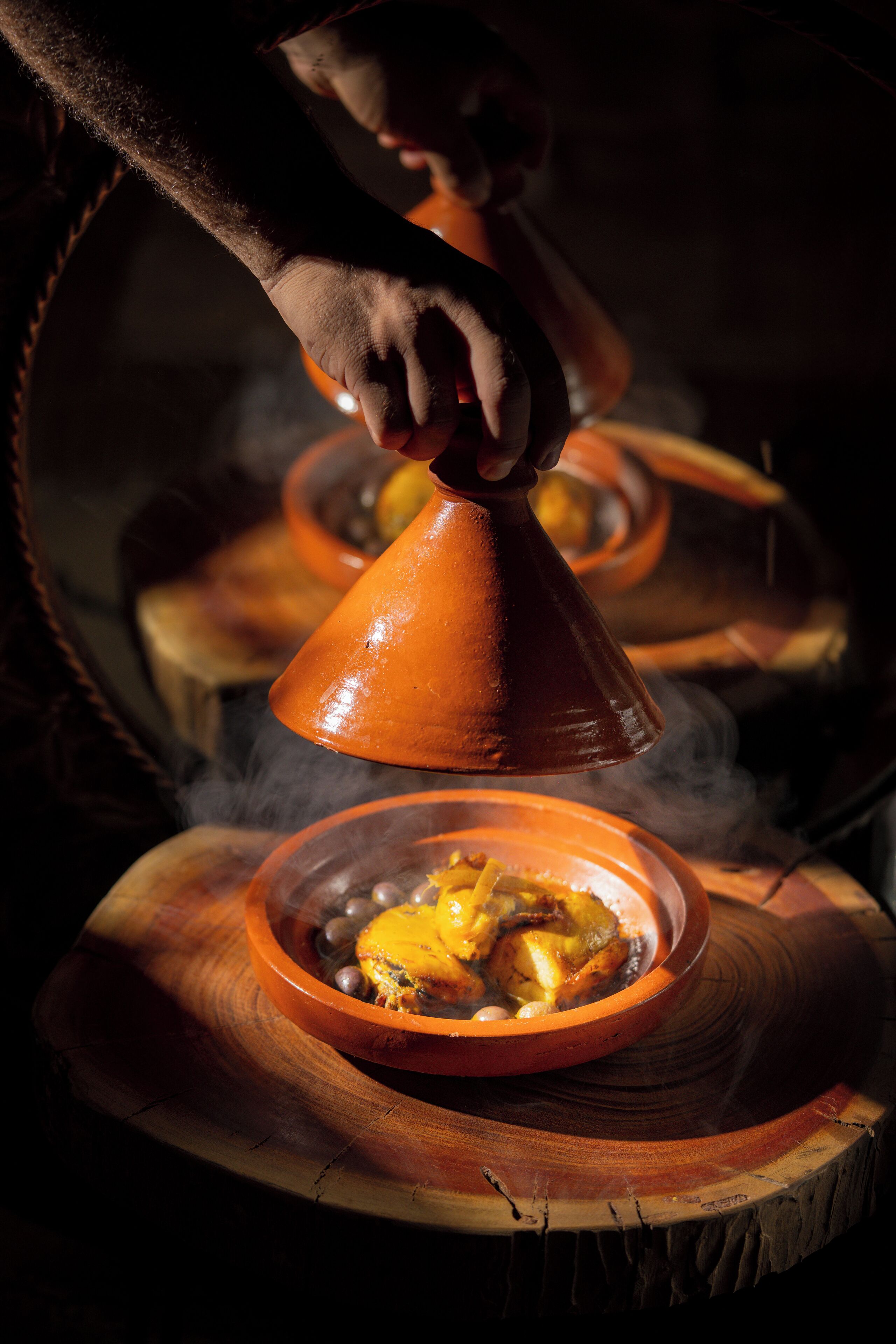 A hand pouring a liquid from a conical vessel over a dish containing food, with steam rising from the dish.