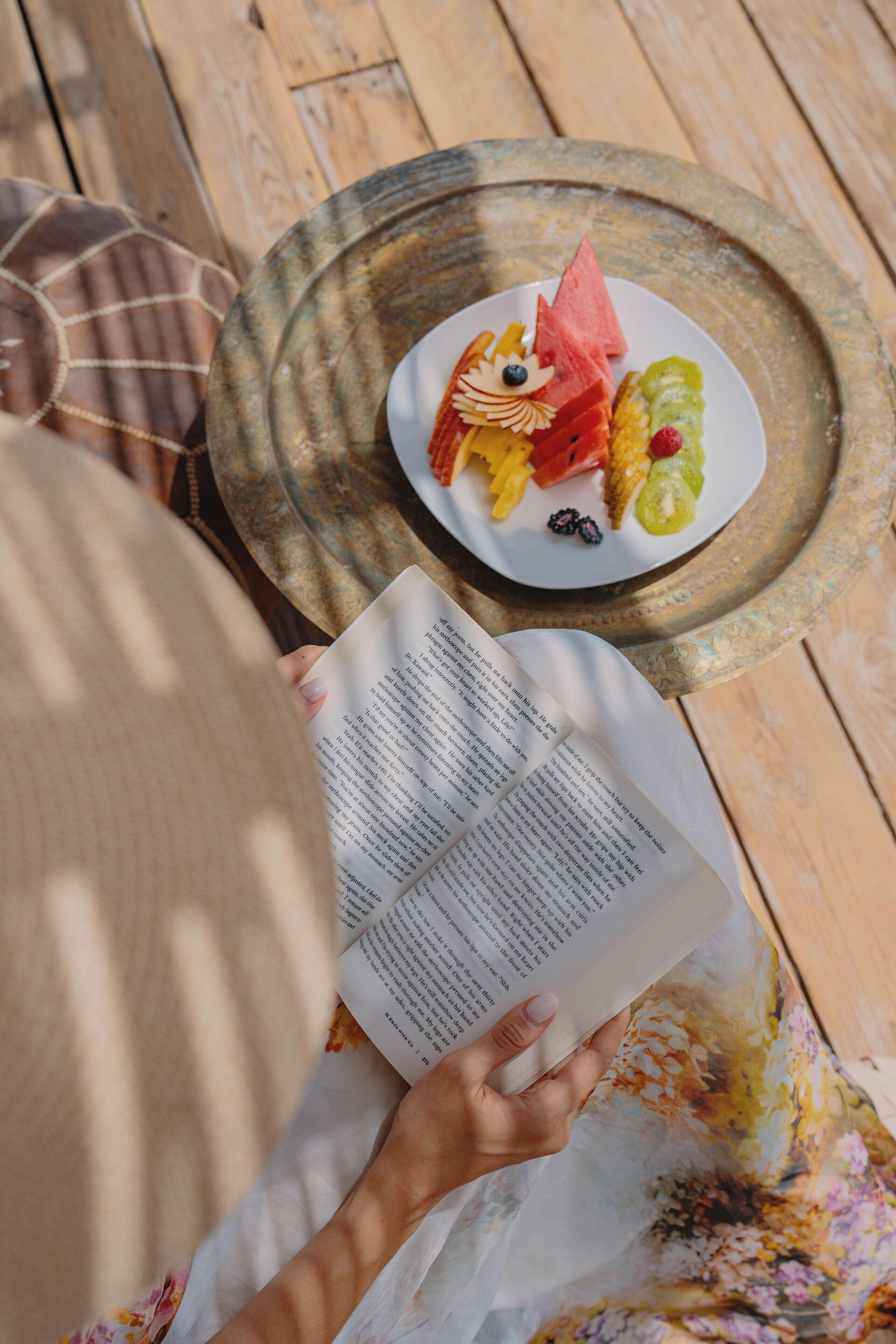 Une personne est assise à une table en bois, un livre ouvert devant elle et une assiette de fruits à portée de main.