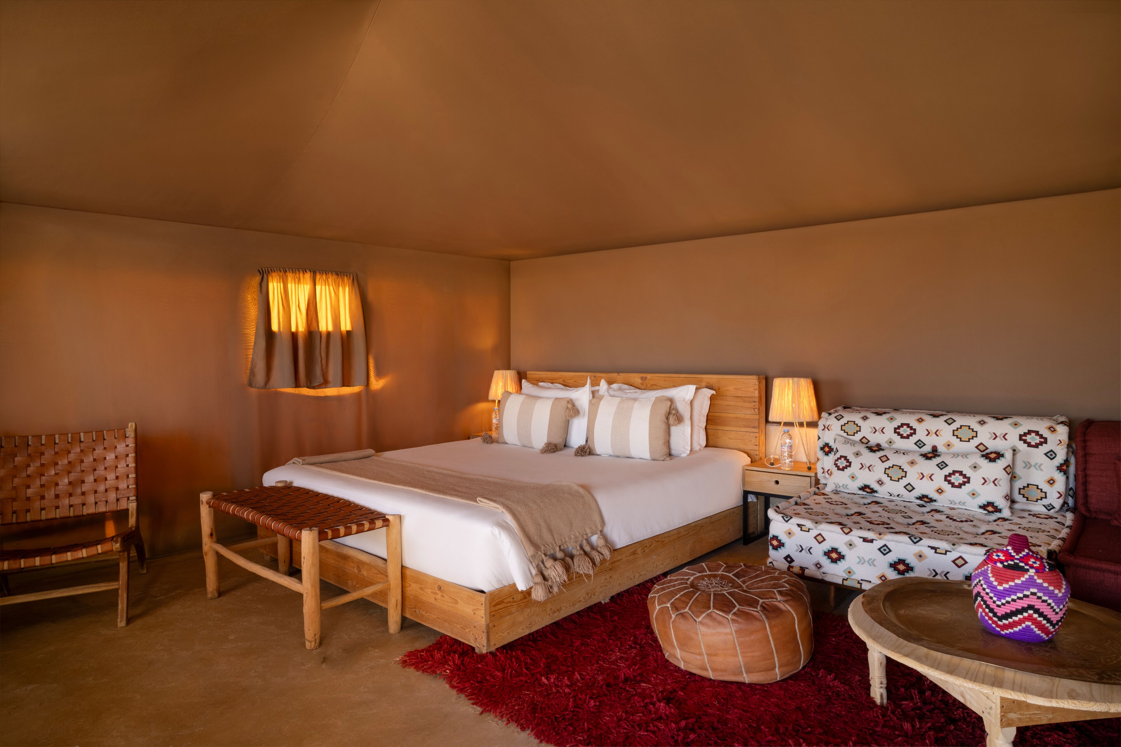 A bedroom with a wooden bed, a red rug, a wooden bench, a patterned sofa, and a small table with a decorative item.