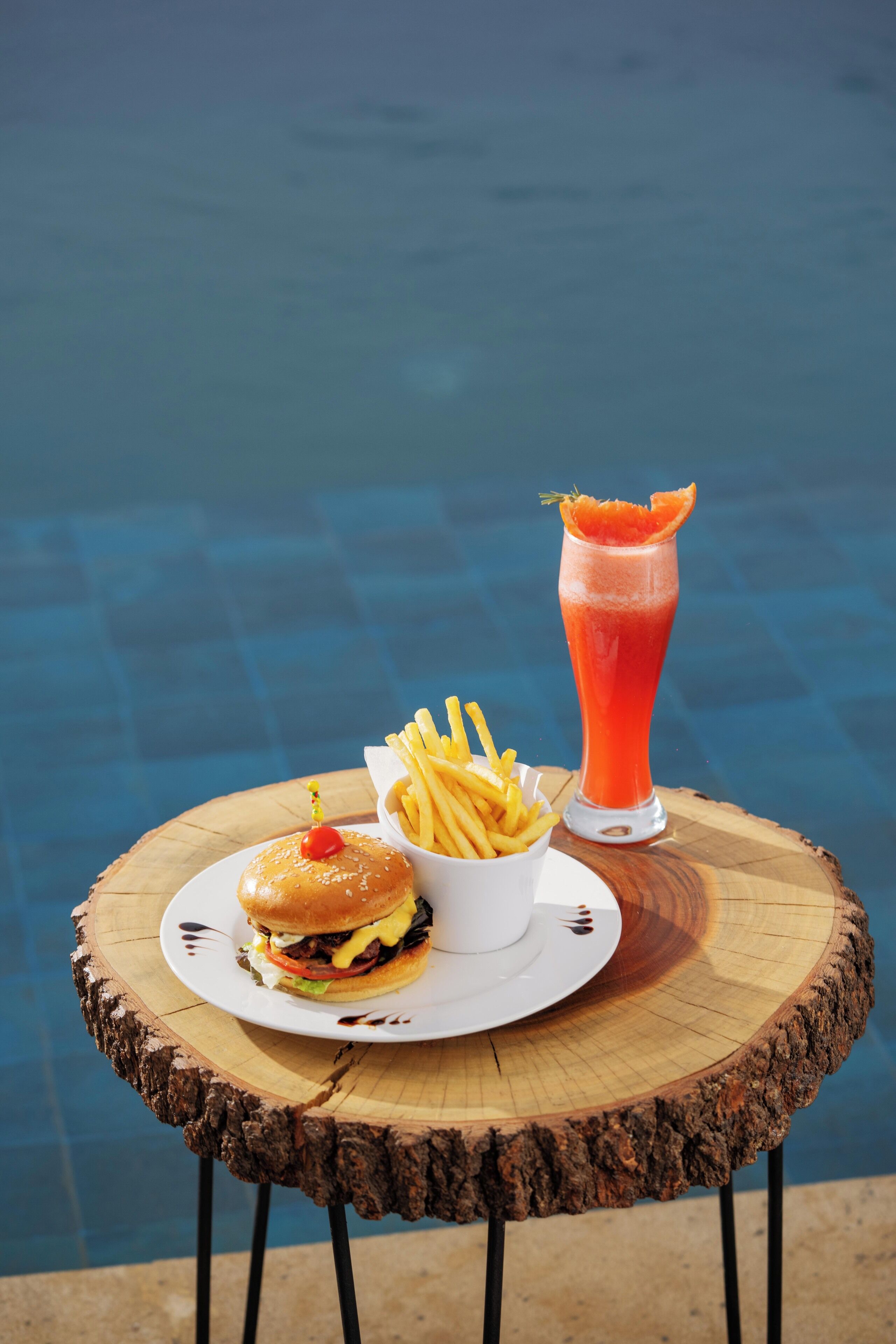 Uma refeição com hambúrguer, batatas fritas e bebida sobre uma mesa de madeira junto a uma piscina.