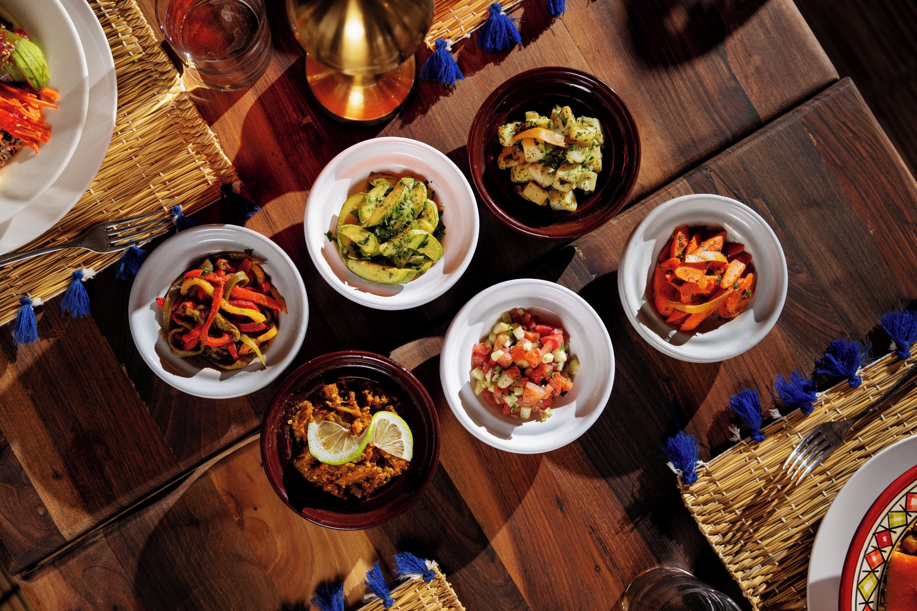 A wooden table with six bowls of food, a candle, and blue tassels.