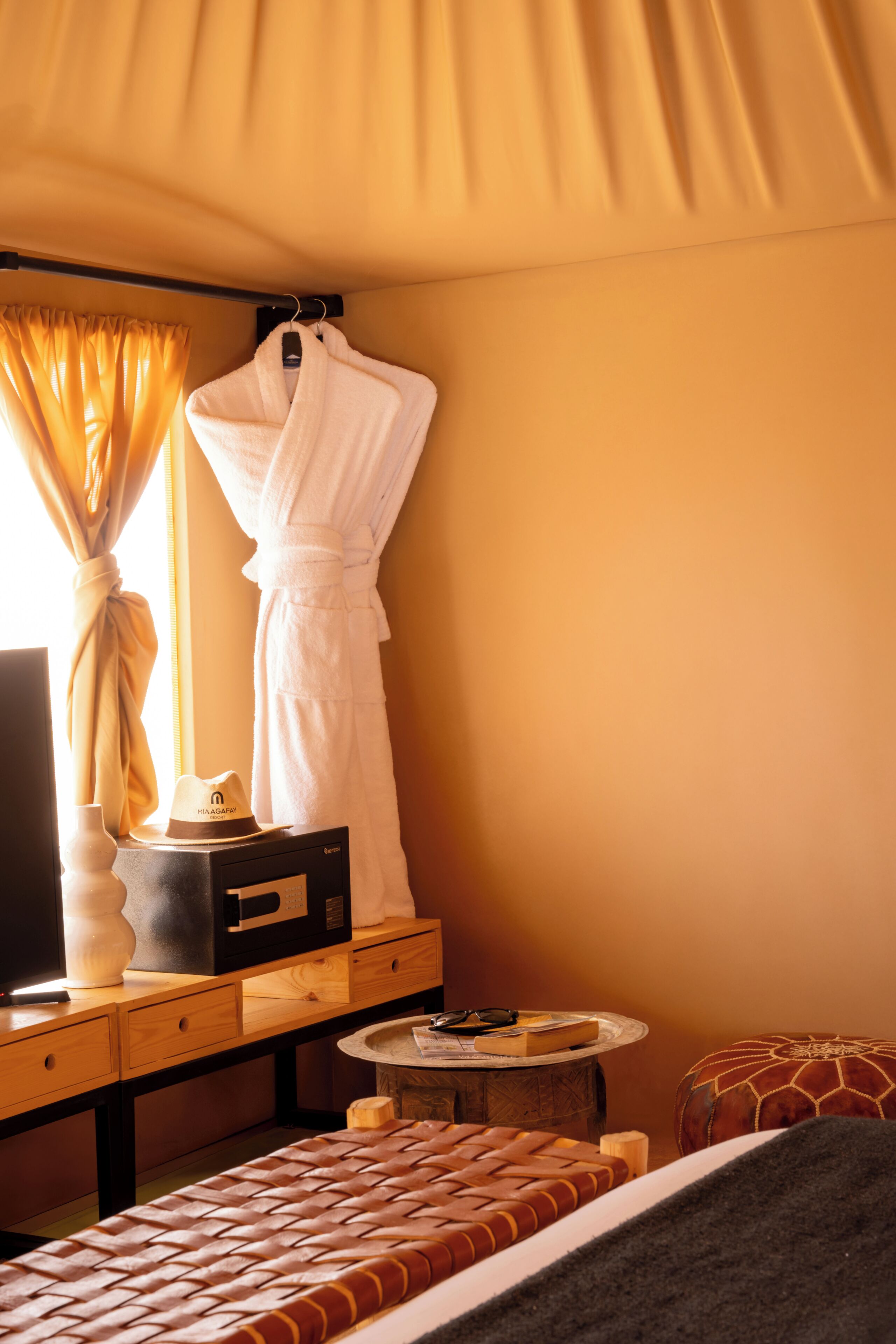 Une chambre d’hôtel avec un lit, un peignoir, un chapeau, une petite table d’appoint et une petite table ronde.