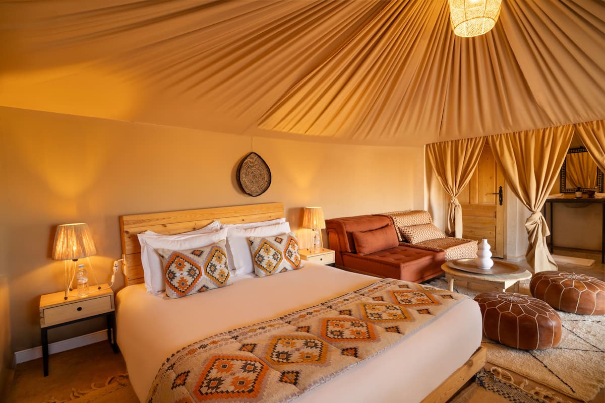 A room with a bed, nightstands, a chair, ottomans, and a lamp.