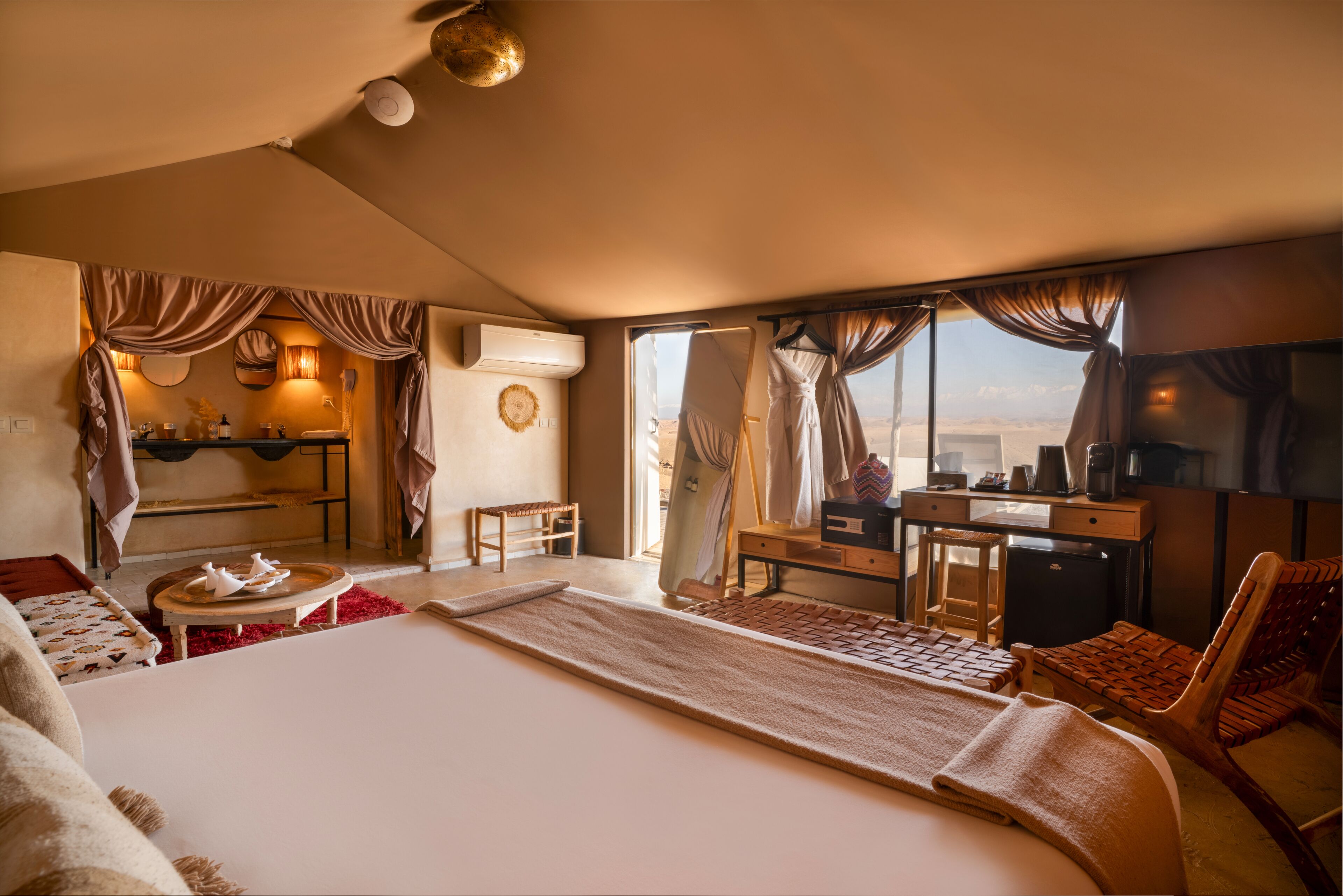 A hotel room with a large bed, a desk, a chair, a small table, and a view of the desert.