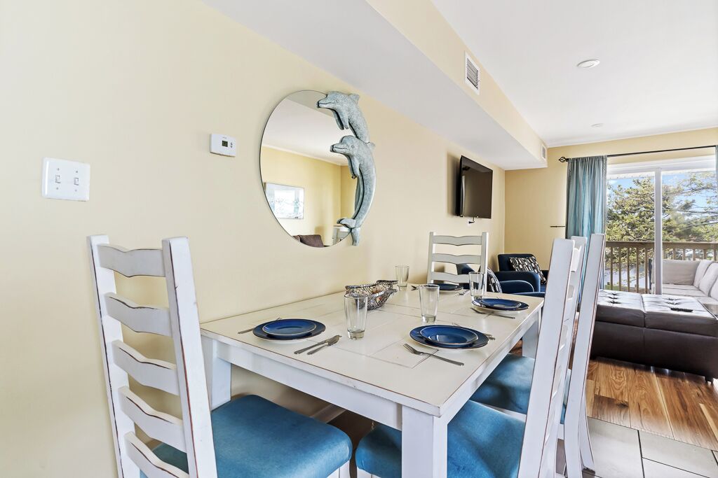 A dining area with a white table set for four, blue chairs, a round mirror with a dolphin sculpture, and a TV mounted on the wall.
