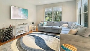 A living room with a grey sofa, a coffee table, a TV mounted on the wall, and a large window with blinds. - Relax and recharge in comfort at this stylish Heritage Village retreat! (Lewes)