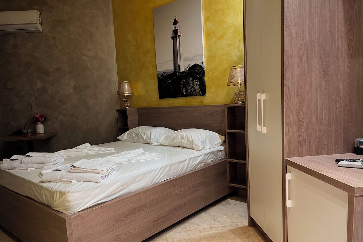 Ein ordentlich bezogenes Bett mit weißen Bettwäsche und Handtüchern, ein Holz-Kopfteil, ein Nachttisch mit Lampe, eine Wand-Klimaanlage und ein Gemälde eines Leuchtturms.