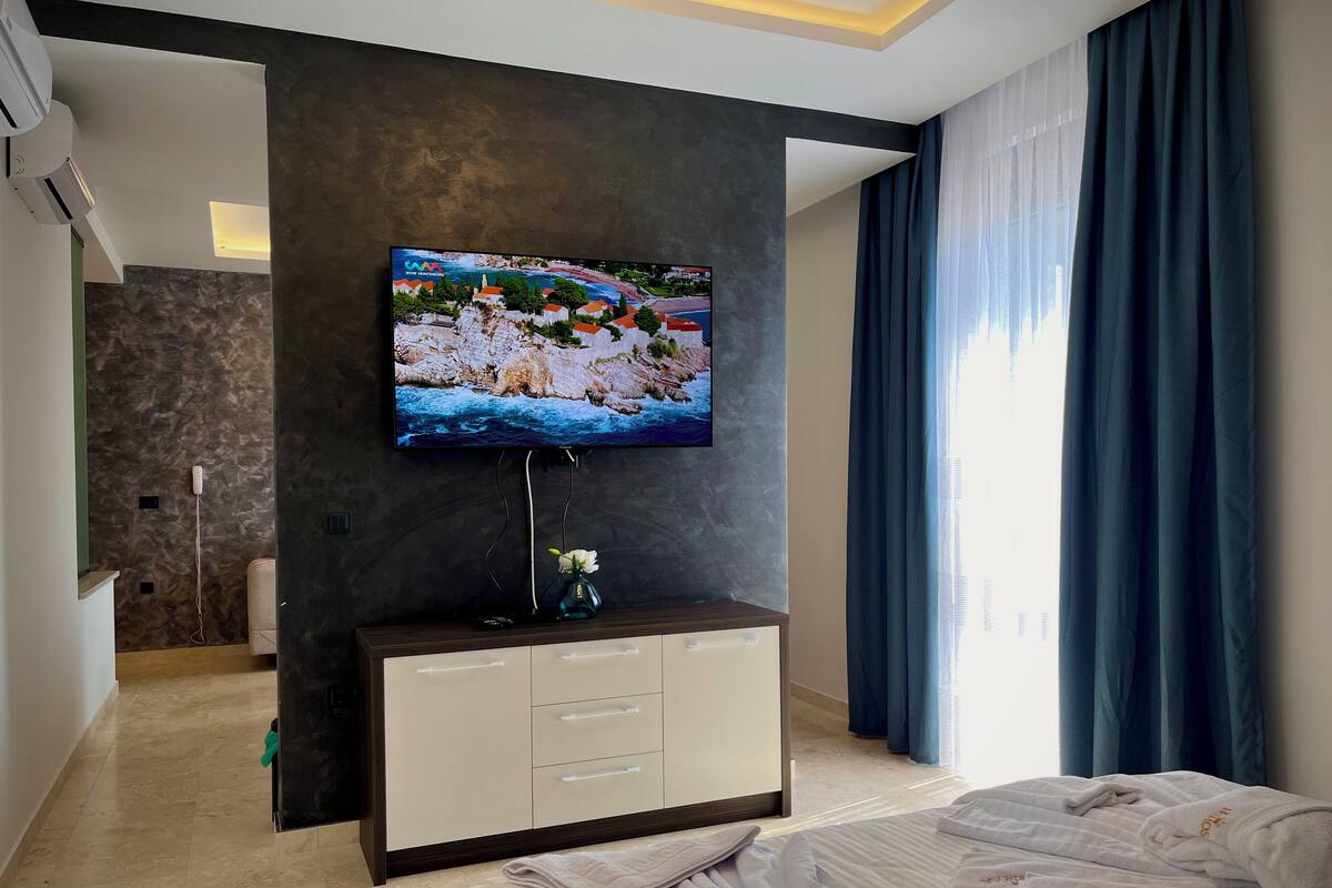Ein modernes Hotelzimmer mit einem an einer dunklen Wand montierten Flachbildfernseher, einer Minibar, einem Bett mit ordentlich gefalteten weißen Handtüchern und einem großen Fenster mit blauen Vorhängen.