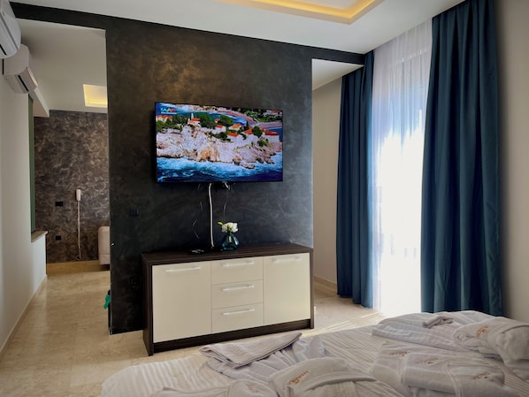A modern hotel room with a flat-screen TV mounted on a dark wall, a minibar, a bed with neatly folded white towels, and a large window with blue curtains.