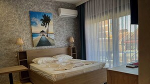 Une chambre d’hôtel avec un lit en bois, deux tables de chevet avec des lampes, un tableau représentant une scène de plage et une vue à travers des rideaux transparents.