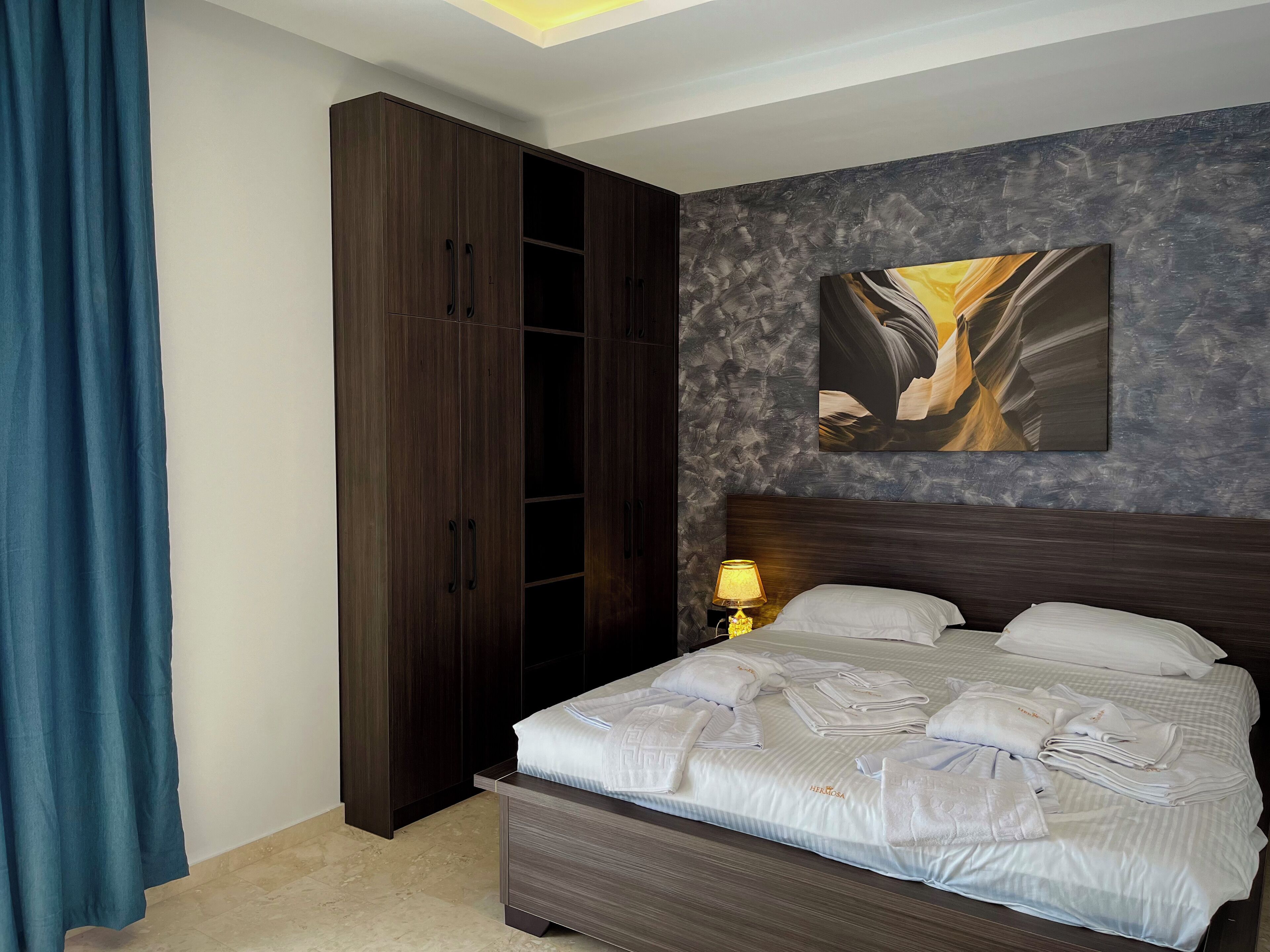 Habitación de hotel con una cama grande, un armario, un cuadro en la pared y una mesita de noche con una lámpara.