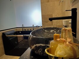 Uma casa de banho moderna com uma grande pia de mármore preto, bancada preta e um pequeno recipiente dourado com produtos de higiene pessoal.