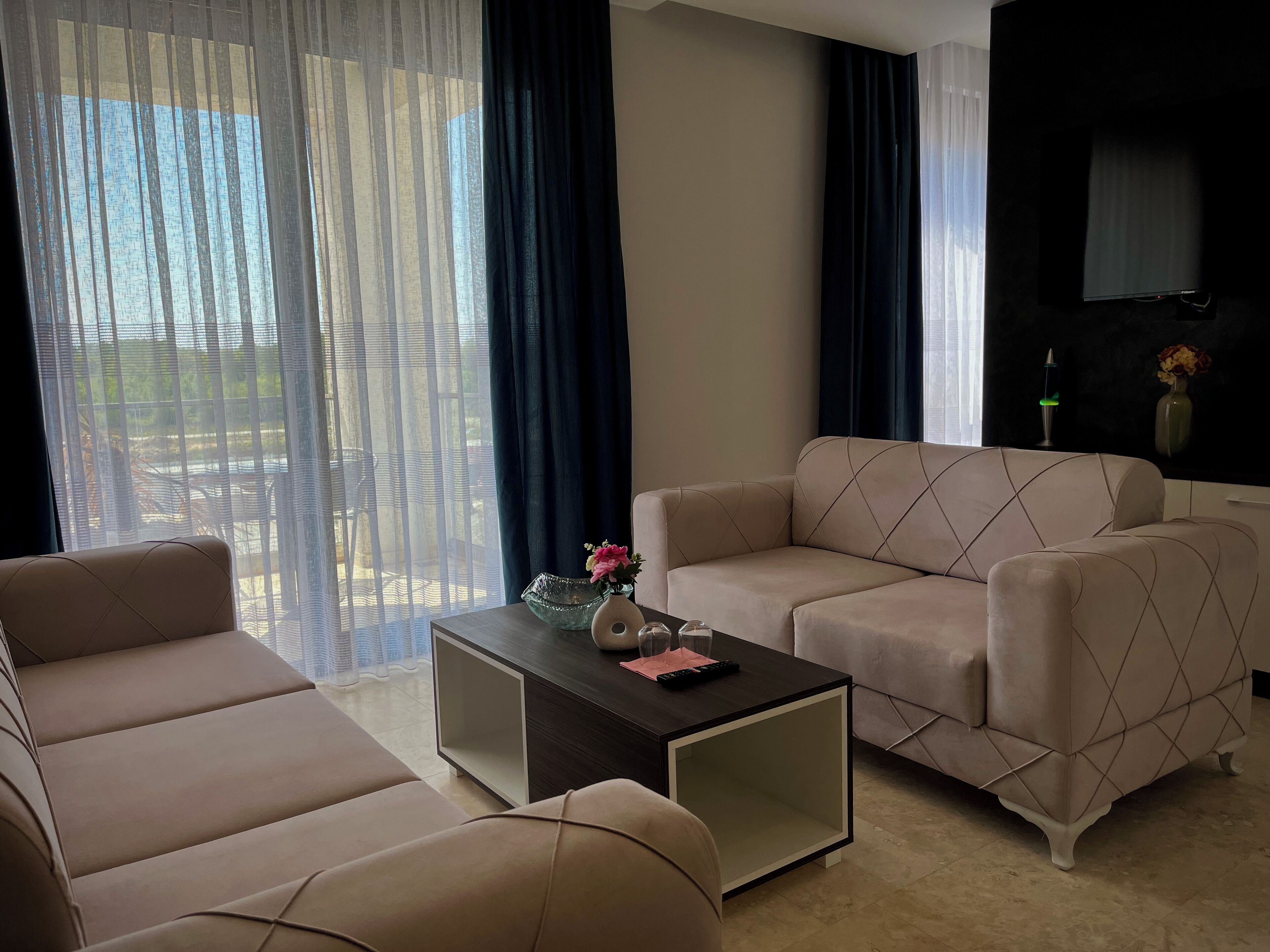 Una sala de estar moderna con un sofá beige, una mesa de centro oscura con adornos, y vistas al exterior a través de cortinas traslúcidas.
